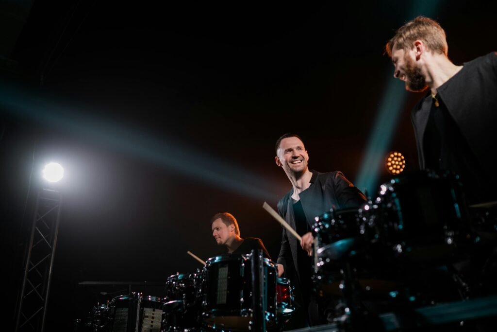 Three male drummers perform energetically on stage under dramatic lighting.
