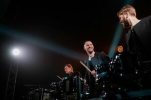 Three male drummers perform energetically on stage under dramatic lighting.